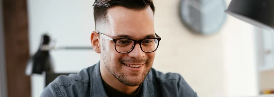 Homem branco, e de óculos grau sorri enquanto olha para a tela do computador. Veste uma camiseta preta com uma camisa social verde sobreposta.