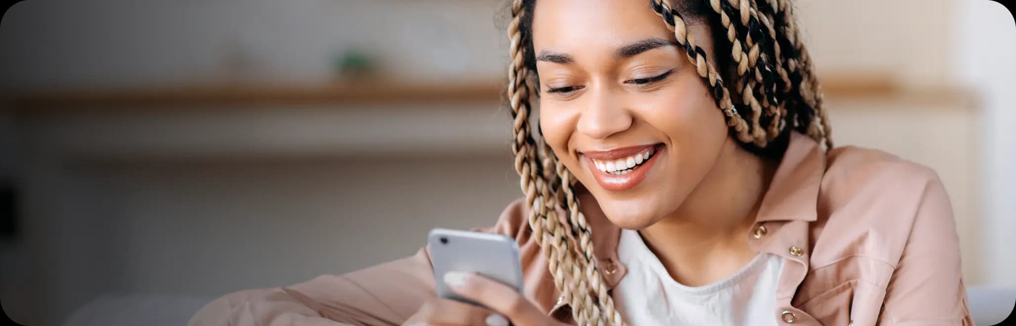 Mulher negra de tranças está sorrindo para o celular enquanto o segura com as duas mãos. Ela usa uma camiseta branca e camisa aberta da cor rosa claro. 