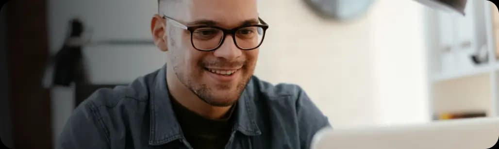 Homem branco de cabelo curto e escuro sorri enquanto utiliza o Asaas em seu notebook. Ele usa óculos de grau e está vestindo camiseta jeans azul.