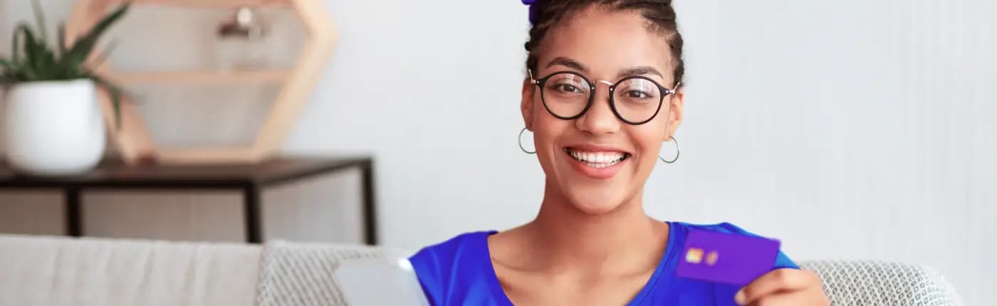 Mulher sorri para a câmera enquanto segura um cartão de crédito na mão esquerda. Ela está de cabelos presos, veste uma camisa azul, além de usar óculos de armação preta e brincos de argola.
