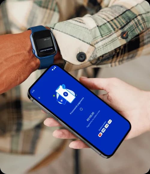 Pessoa usando um relógio inteligente e aproximando seu punho a um celular que está na mão de outra pessoa. Esse celular exibe em sua tela o processamento do pagamento, enquanto o relógio exibe uma imagem do cartão virtual.