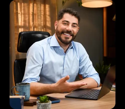 Evaristo Costa sentado em uma mesa com o notebook em sua frente. Ele aponta para a tela e sorri.