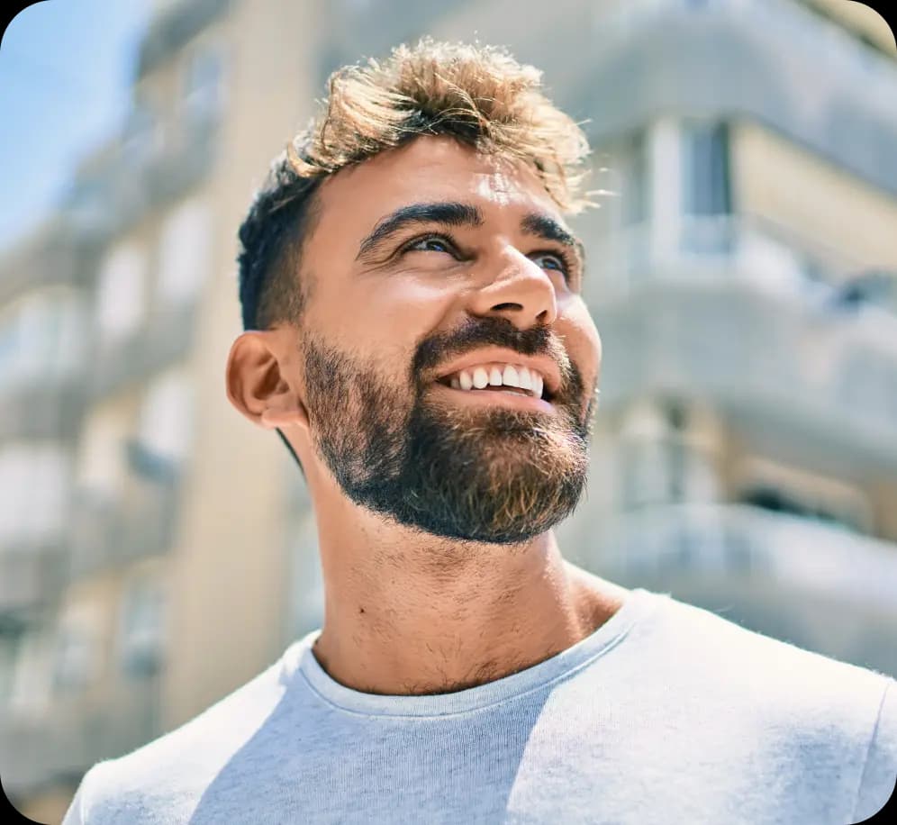 Empreendedor olha para cima, sorrindo. Ele tem a pele clara e cabelos castanhos, usa barba e veste uma camiseta na cor cinza. Ao fundo, há um prédio em desfoque.
