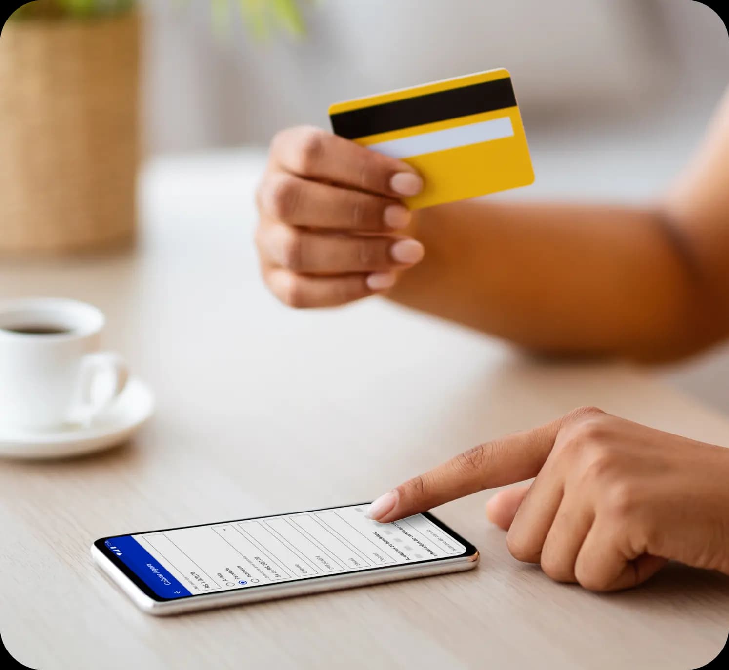 A mão direita de uma pessoa segura um cartão de crédito, enquanto sua mão esquerda gera uma cobrança no aplicativo do Asaas. Ao fundo, em desfoque, há uma xícara de café e uma planta.
