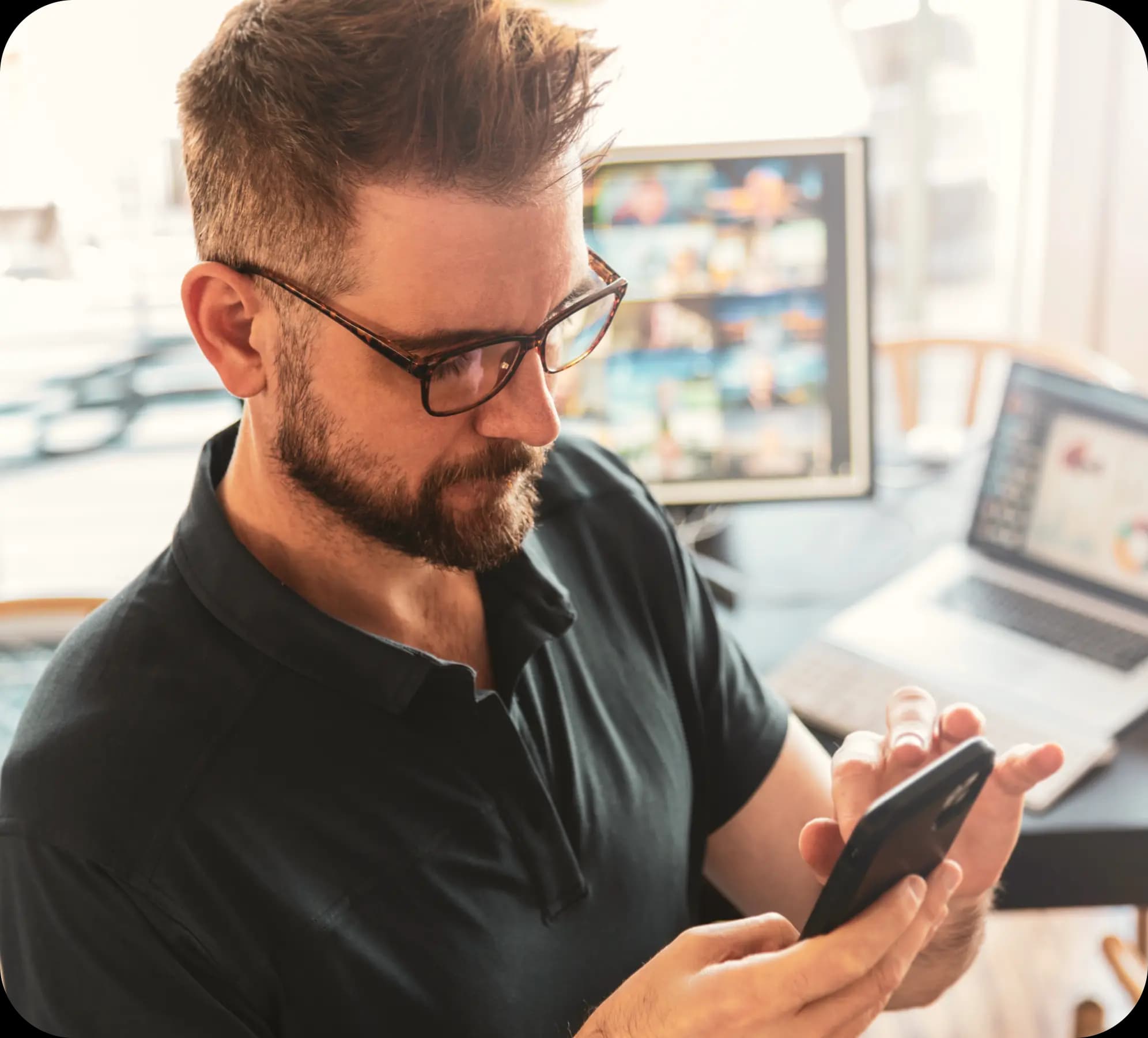 Empreendedor mexe no celular, enquanto faz uma expressão séria, indicando que está concentrado. Ele tem cabelos castanhos e pele clara, e veste uma camisa preta e óculos de armação preta.