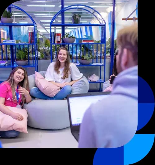 Colaboradores do Asaas conversam na área criativa. Há duas mulheres sorrindo, uma está sentada em um puff e a outra de joelhos no chão, com uma almofada no colo. No canto inferior direito, há um homem de costas, em desfoque. Ao fundo, estão duas estantes coloridas em azul e algumas plantas.